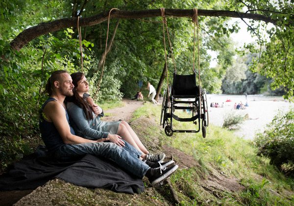 junger Mann und junge Frau in der Natur sitzend, über Ihnen ein Rollstuhl, der an einer Schnur an einem Ast hängt