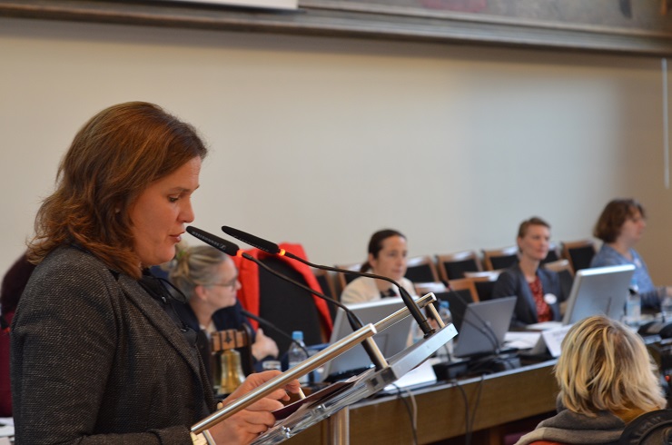 Eine Frau redet in ein Mikrofon. Im Hintergrund sind unscharf Personen mit Laptops zu sehen.