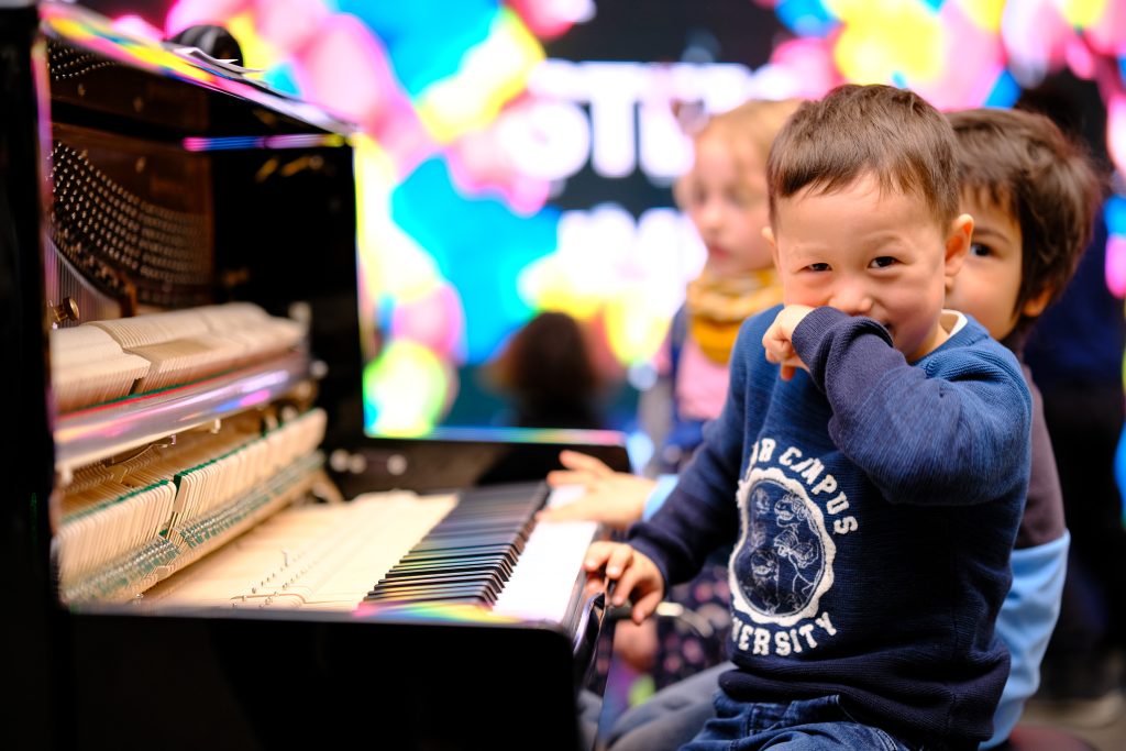 Zwei Jungen im Grundschulalter sitzen an einem Klavier. Beide schauen in die Kamera. Der vordere Junge lacht und hält sich seine Hand vor den Mund.