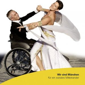 Kartenausschnitt: Ein Mann und eine Frau tanzen zusammen. Der Mann trägt einen schwarzen Anzug. Die Frau trägt ein weißes Kleid mit goldenen Verzierungen. Es sieht wie eine Tanz-Aufführung aus. Der Mann ist im Rollstuhl.