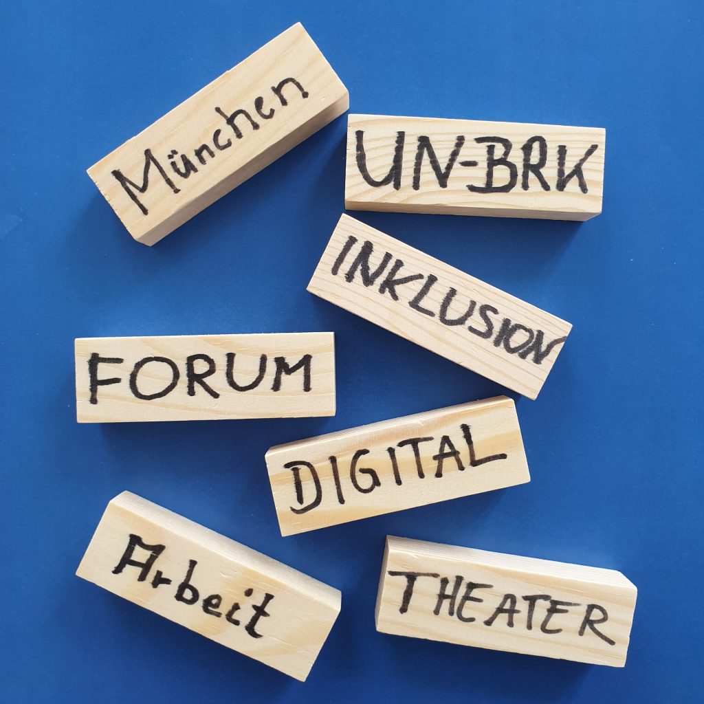 Auf dem Foto sieht man kleine Holzklötzchen, die verstreut auf einer blauen Fläche liegen. Jedes Klötzchen ist mit einem anderem Wort beschriftet. Dies sind die Wörter: "München", "UN-BRK", "Inklusion", "Forum", "Digital", "Arbeit" und "Theater".