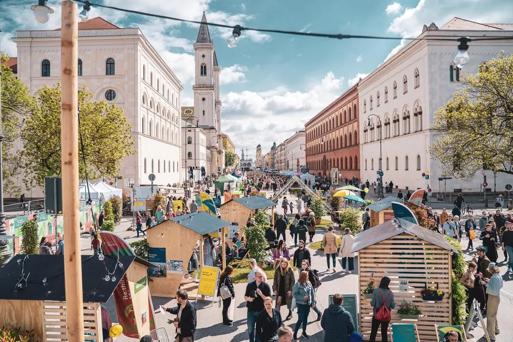 Auf dem Foto sieht man die verkehrsfreie Ludwigstraße. Auf der Festival-Meile stehen viele Holzhütten und Grünpflanzen. Dazwischen sind viele Menschen zu Fuß unterwegs.