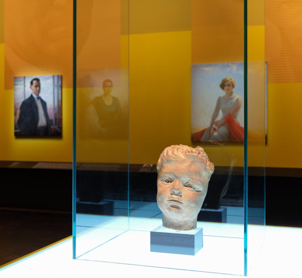 Das Foto zeigt eine Ausstellung im Jüdischen Museum München. Vorne rechts ist eine Büste von einem Kind zu sehen. Dahinter hängen zwei Portraits an einer großen gelben Wand.
