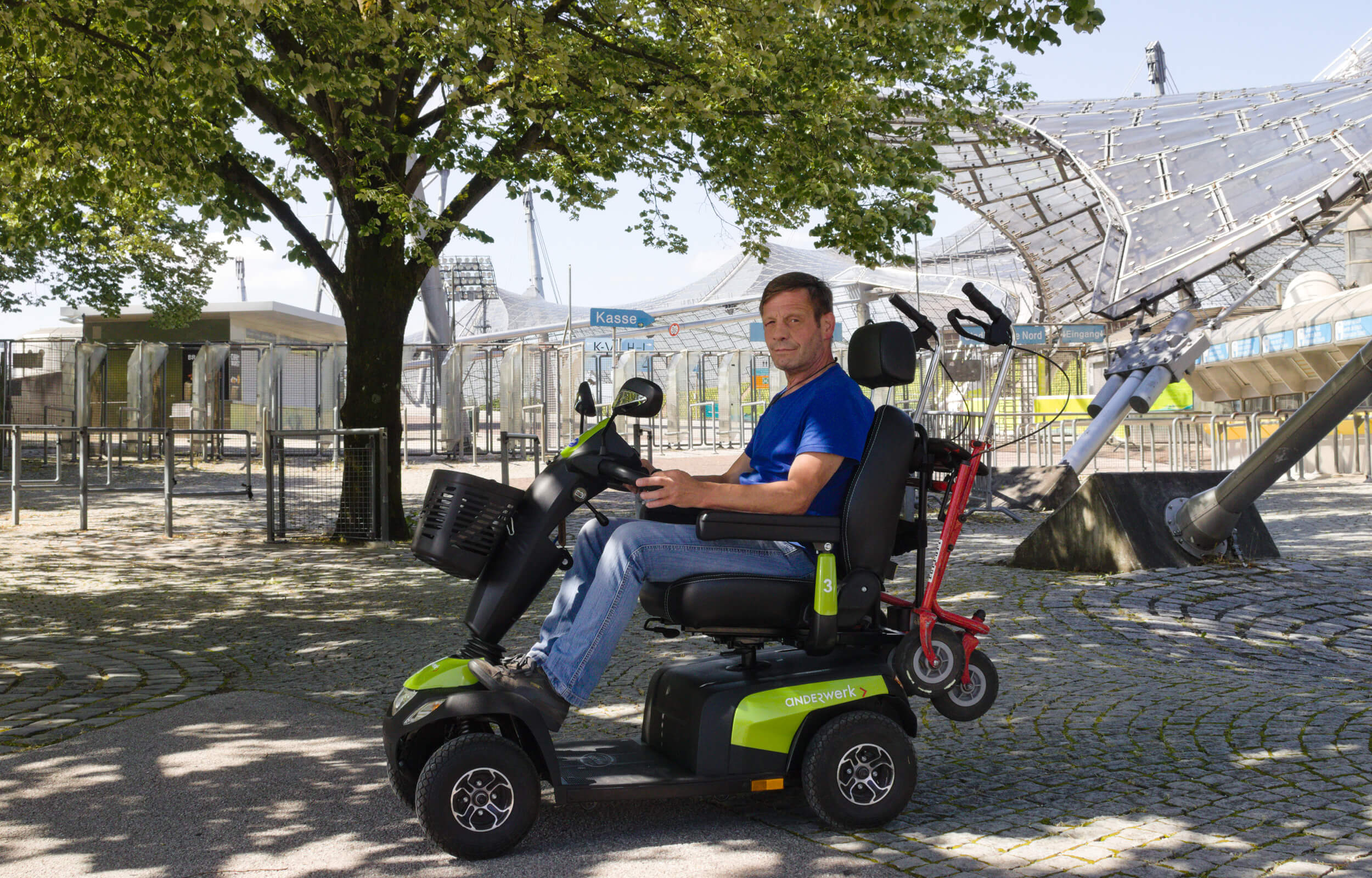 Ein Mann sitzt in einem Elektromobil. Auf dem Elektromobil steht "anderwerk". Im Hintergrund ist das Zeltdach des Olympiaparks