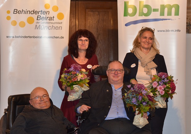 Vier Menschen sitzen und stehen vor zwei Roll-Ups und lächeln. Auf dem linken Roll-Up steht Behindertenbeirat der Landeshauptstadt München www.behindertenbeirat-muenchen.de. Auf dem rechten Roll-Up steht bb-m www.bb-m.info.