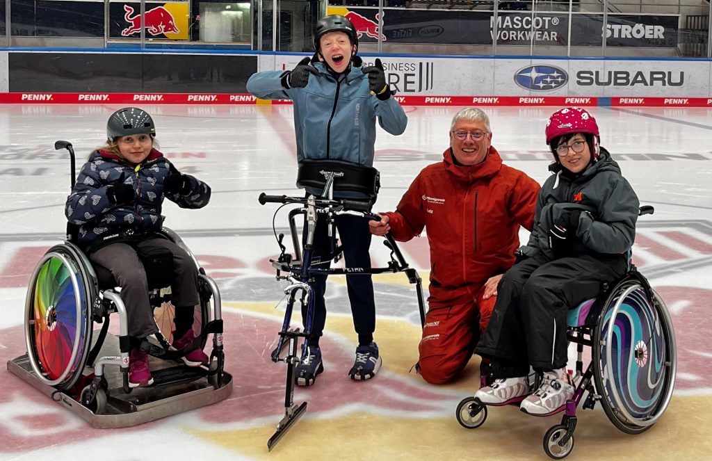 Das Foto zeigt vier Menschen nebeneinander auf einer Eisfläche in einer Halle. Ein Mädchen sitzt im Rollstuhl. Deren Rollstuhlräder stecken in einem Rollstuhl-Gleiter. Ein Junge steht in einem Ice Frame-Runner. Ein weiteres Mädchen sitzt in einem Rollstuhl. Daneben kniet ein Mann.