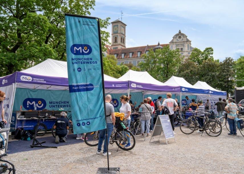 Auf dem Foto sieht man mehrere Pavillons im Freien. Davor steht eine Fahne mit dem Logo "München unterwegs" und Menschen mit Fahrrädern.