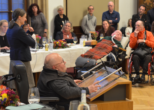 Foto: Der Behindertenbeauftragte Oswald Utz sitzt in seinem Rollstuhl vor dem Rednerpult im Sitzungssaal des Münchner Rathauses. Links von ihm gebärdet eine Gebärdensprachdolmetscherin. Dahinter und davor hören Menschen mit und ohne Behinderungen zu.